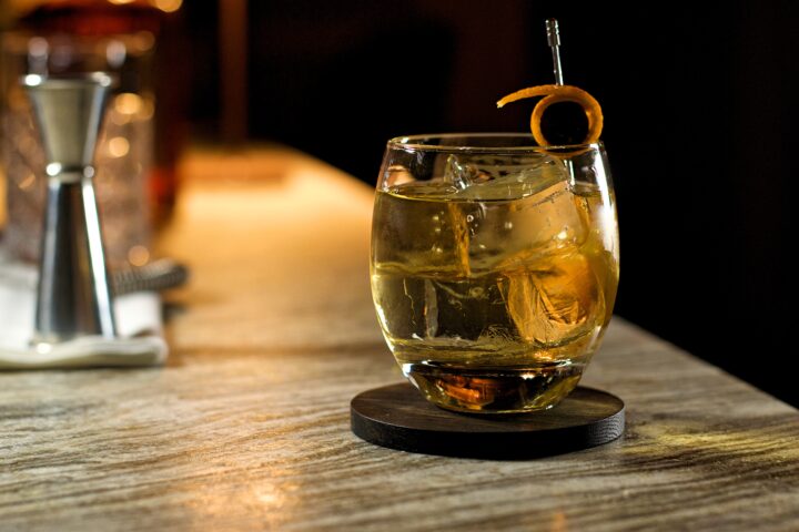 bicchiere tondo con ghiaccio gristallino e un cocktail con vodka e amaretto, sullo sfondo un jigger e una bottiglia di disaronno